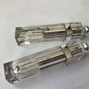 May include: Two clear, rectangular cosmetic containers with silver accents and a rounded silver cap. The text "Beyond Beauty by Jennifer" is printed on the side of each container. The containers are on a white surface.