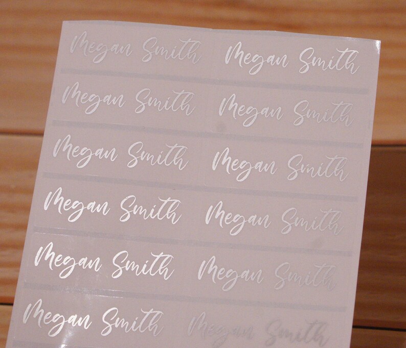 May include: A sheet of rectangular stickers with the name "Megan Smith" printed in a cursive font. The text is in a silver color, and the stickers are arranged in rows and columns.