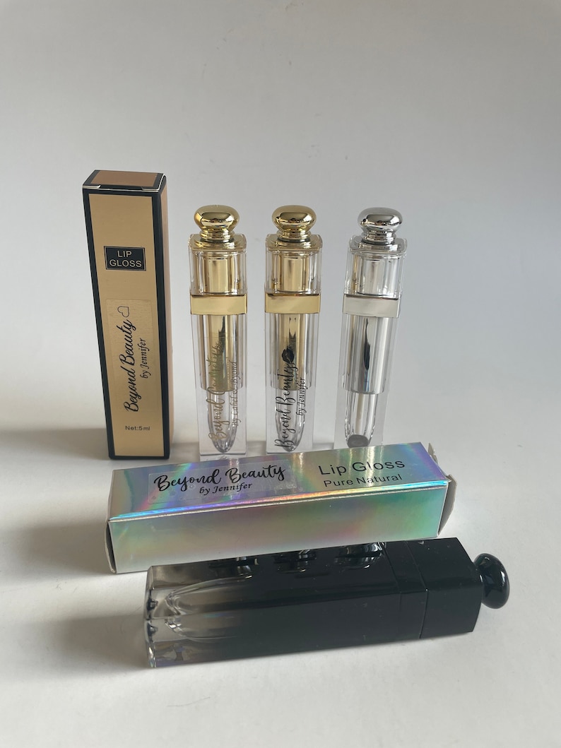 May include: Lip gloss products by Beyond Beauty by Jennifer. The image shows three clear lip gloss tubes with gold and silver caps, and two boxes. One box is black and the other is holographic.