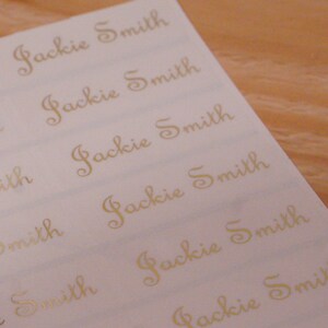 May include: A sheet of white labels with the name "Jackie Smith" printed in gold cursive script. The labels are arranged in rows and columns, with a wooden surface in the background.
