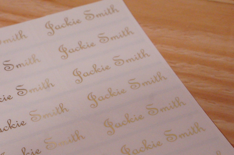 May include: A sheet of white labels with the name "Jackie Smith" printed in gold cursive script. The labels are arranged in rows and columns, with a wooden surface in the background.