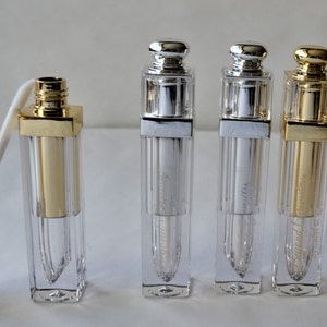 May include: A collection of clear, rectangular lip gloss tubes with gold and silver-colored caps. One tube has a white applicator extending from it. The tubes have a gold or silver metallic finish and are labeled with text.