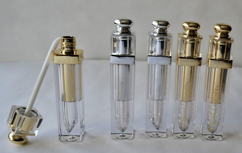May include: A collection of clear, rectangular lip gloss tubes with gold and silver-colored caps. One tube has a white applicator extending from it. The tubes have a gold or silver metallic finish and are labeled with text.