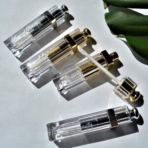 May include: Four clear lip gloss tubes with silver and gold accents are arranged on a white surface. The tubes have text that reads "Beyond Beauty" and "by Jennifer" or "by Label Diamond". One tube has the applicator extended.