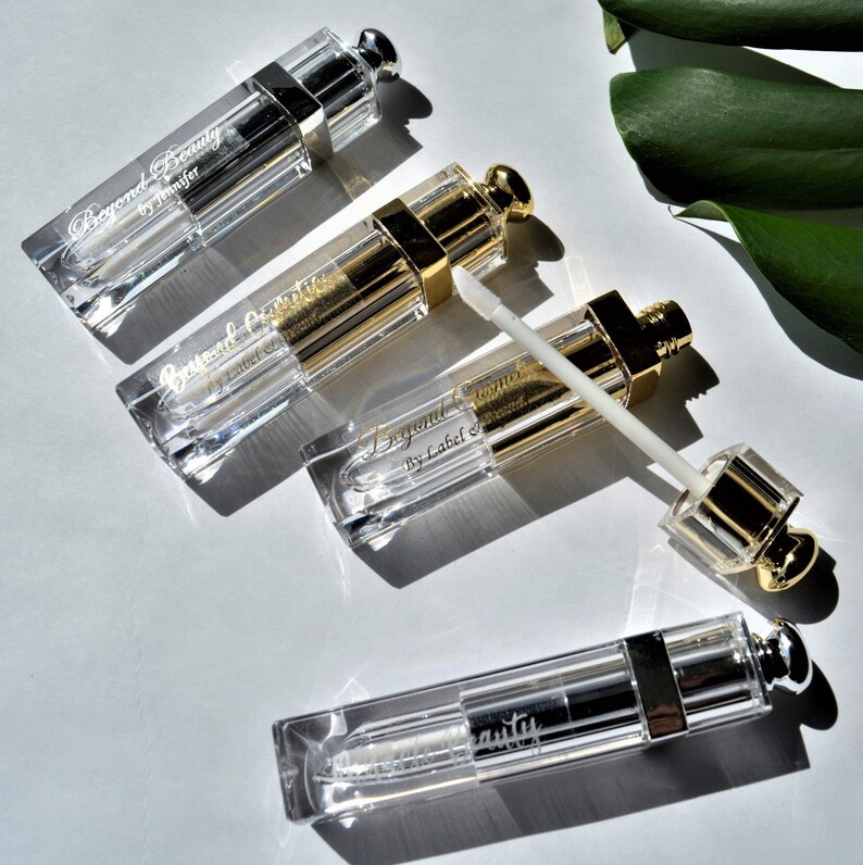 May include: Four clear lip gloss tubes with silver and gold accents are arranged on a white surface. The tubes have text that reads "Beyond Beauty" and "by Jennifer" or "by Label Diamond". One tube has the applicator extended.