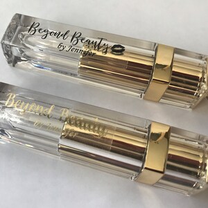 May include: Two clear lipstick tubes with gold accents. The tubes have a rectangular shape with a gold band and a rounded gold cap. The text "Beyond Beauty by Jennifer" is printed on the tubes.