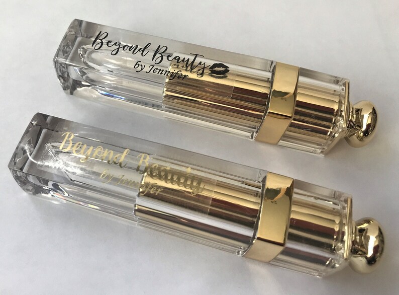May include: Two clear lipstick tubes with gold accents. The tubes have a rectangular shape with a gold band and a rounded gold cap. The text "Beyond Beauty by Jennifer" is printed on the tubes.