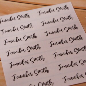 May include: A sheet of white labels printed with the name "Jennifer Smith" in a black, elegant script font. The labels are arranged in rows and columns, ready for use. The background is a light wood surface.