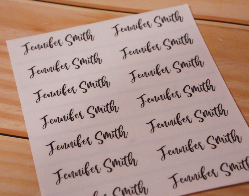 May include: A sheet of white labels printed with the name "Jennifer Smith" in a black, elegant script font. The labels are arranged in rows and columns, ready for use. The background is a light wood surface.