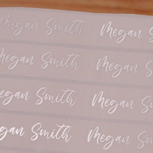 May include: A sheet of white labels with the name "Megan Smith" printed in a cursive font. The labels are arranged in rows and columns, with the name repeated multiple times. The background is a light pink color.