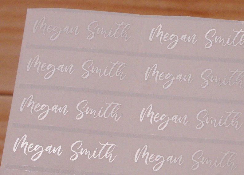 May include: A sheet of white labels with the name "Megan Smith" printed in a cursive font. The labels are arranged in rows and columns, with the name repeated multiple times. The background is a light pink color.