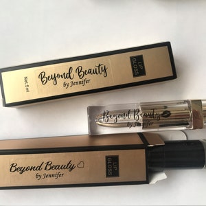 May include: Lip gloss products from Beyond Beauty by Jennifer. The image shows two boxes and a lip gloss tube. The boxes are gold and black with the text "Beyond Beauty by Jennifer" and "Lip Gloss". The lip gloss tube is clear with a gold cap.