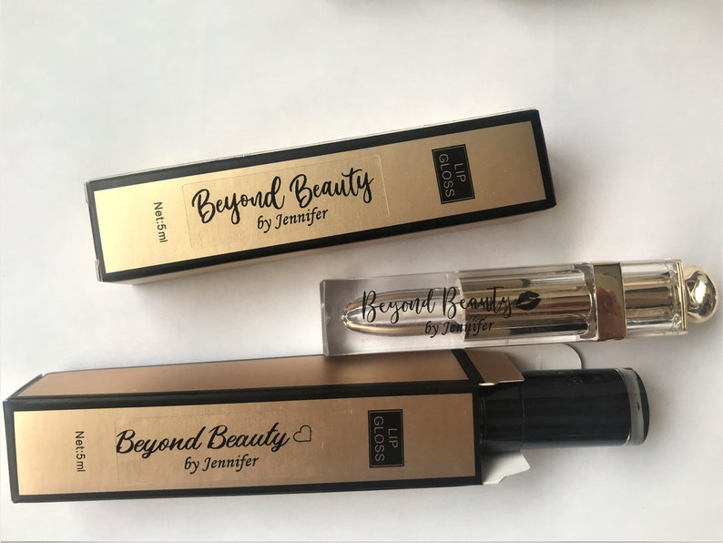 May include: Lip gloss products from Beyond Beauty by Jennifer. The image shows two boxes and a lip gloss tube. The boxes are gold and black with the text "Beyond Beauty by Jennifer" and "Lip Gloss". The lip gloss tube is clear with a gold cap.