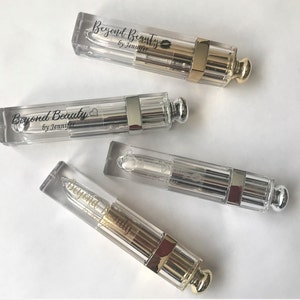 May include: Four clear lip gloss tubes with metallic silver and gold accents. Each tube has the text "Beyond Beauty by Jennifer" printed on it. The lip gloss tubes are arranged diagonally on a white surface.