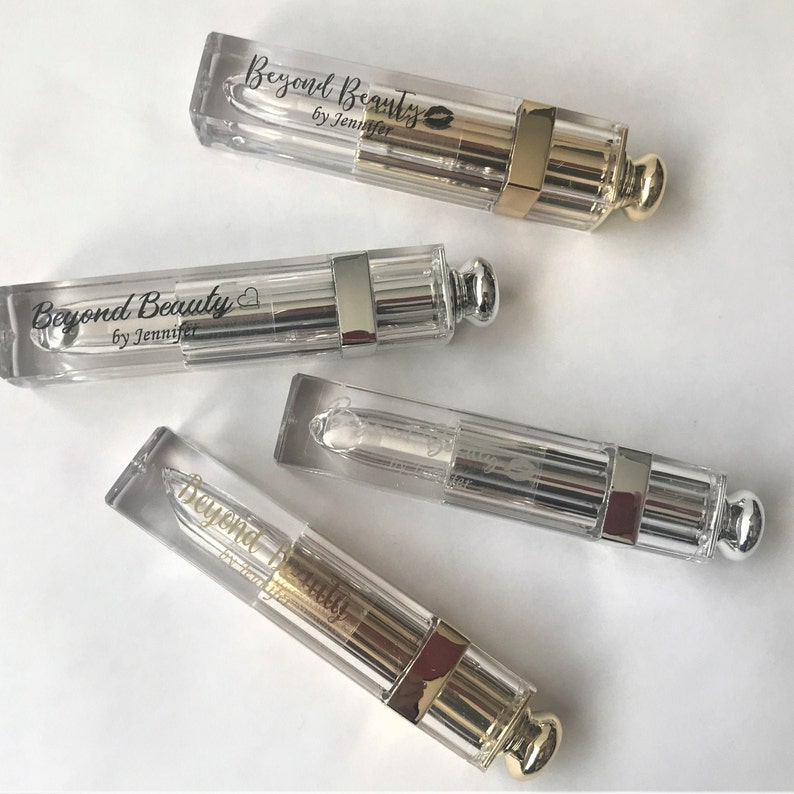 May include: Four clear lip gloss tubes with metallic silver and gold accents. Each tube has the text "Beyond Beauty by Jennifer" printed on it. The lip gloss tubes are arranged diagonally on a white surface.