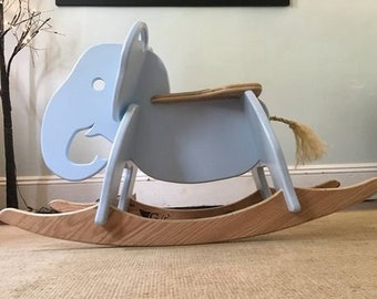 May include: A light blue wooden rocking elephant toy with a natural wood base. The elephant has a saddle and a tail made of rope.
