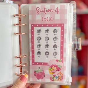 May include: A pink and white planner insert with the text "Section 4 $500". The insert features a grid with berry illustrations and dollar amounts. A Strawberry Shortcake graphic is at the bottom. The insert is held in a rose gold binder.