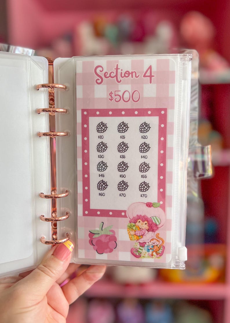 May include: A pink and white planner insert with the text "Section 4 $500". The insert features a grid with berry illustrations and dollar amounts. A Strawberry Shortcake graphic is at the bottom. The insert is held in a rose gold binder.