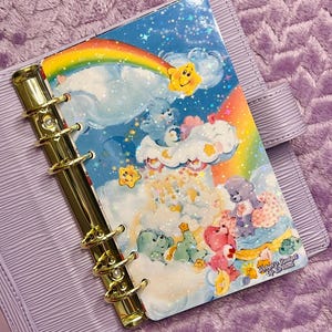 May include: A lilac planner with a gold binder and a colourful insert featuring a rainbow, clouds, and cartoon characters. The insert has the text "Robyn's Budget of Dreams". The planner is on a purple textured surface.
