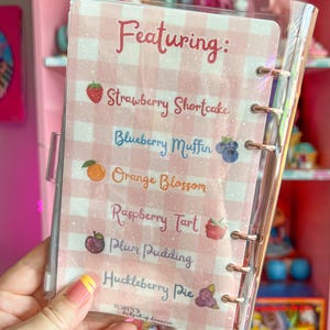 May include: A clear, plastic-covered notebook with a pink and white gingham pattern. The notebook features a list of dessert names, including "Strawberry Shortcake" and "Blueberry Muffin". The text "Featuring:" is at the top.