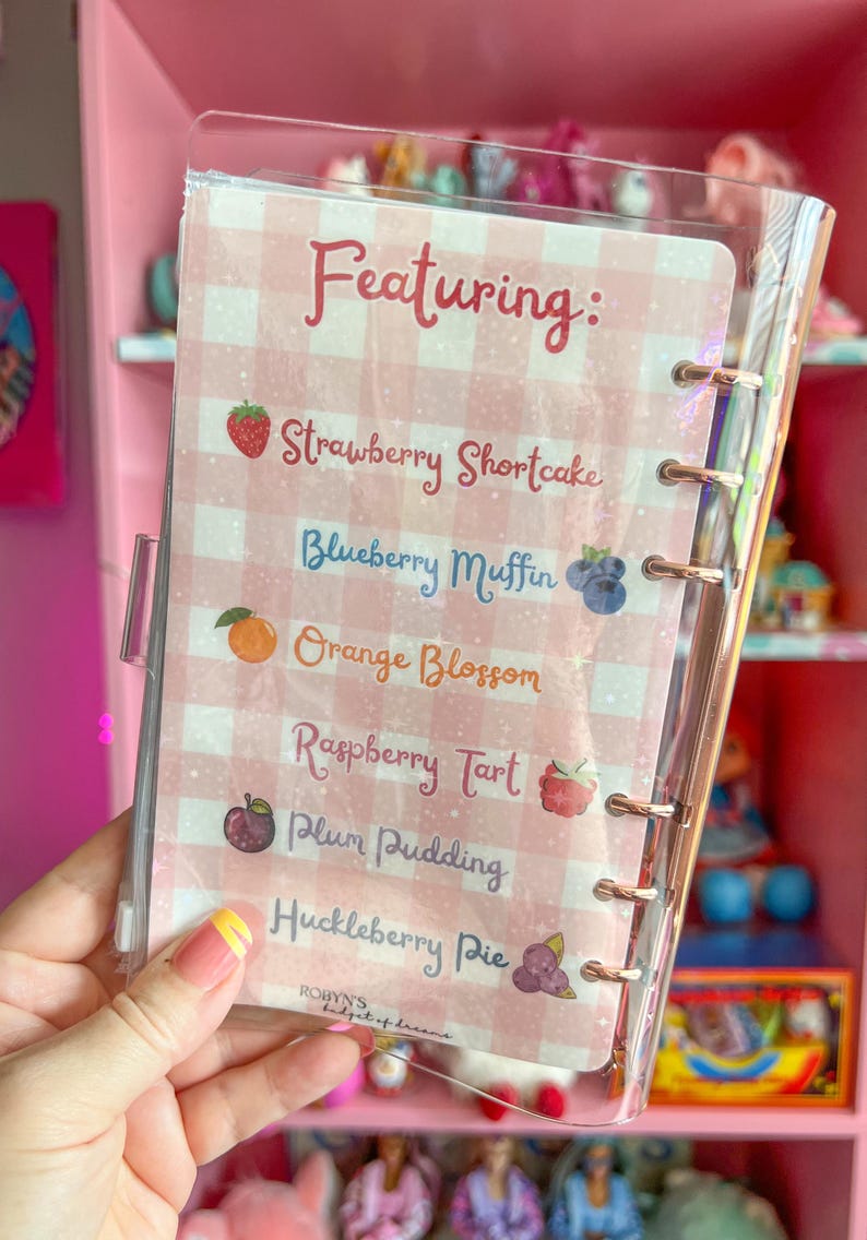 May include: A clear, plastic-covered notebook with a pink and white gingham pattern. The notebook features a list of dessert names, including "Strawberry Shortcake" and "Blueberry Muffin". The text "Featuring:" is at the top.