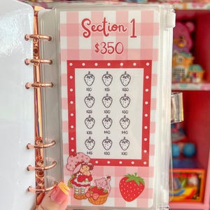 May include: A clear binder with a rose gold spine holding a strawberry-themed savings tracker. The tracker features a pink and white gingham pattern, strawberry illustrations, and the text "Section 1 $350".
