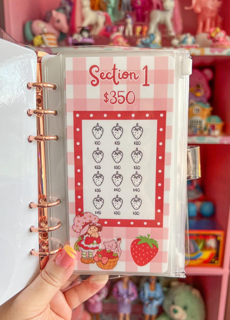 May include: A clear binder with a rose gold spine holding a strawberry-themed savings tracker. The tracker features a pink and white gingham pattern, strawberry illustrations, and the text "Section 1 $350".