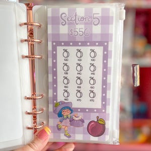 May include: A clear plastic budget binder insert with a purple gingham pattern and the text "Section 5 $550". The insert features illustrations of peaches and a cartoon character. The binder has a rose gold spine.