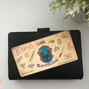 May include: A brown and tan card with a blue and gold Ravenclaw crest and the text "$300 Ravenclaw Robyn's Budget of Dreams Each Icon $20". The card is on a black planner.