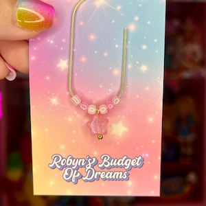 May include: A gold-colored metal bookmark with a pink star charm and pink beads. The bookmark is attached to a card with a gradient background of pink, blue, and yellow. The card has the text "Robyn's Budget Of Dreams".