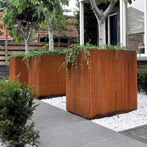 Könnte beinhalten: Drei große, rechteckige, rostige Metall-Pflanzkübel mit grünen Pflanzen, die aus ihnen herauswachsen. Die Pflanzkübel stehen auf einer weißen Kiesfläche.