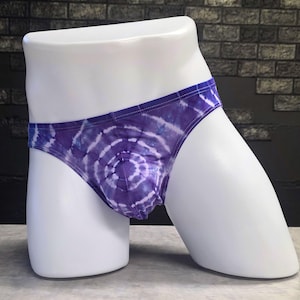 May include: Purple and white tie-dye briefs. The briefs are displayed on a white mannequin. The tie-dye pattern is a spiral design with shades of purple and white. The background is a dark brick wall.