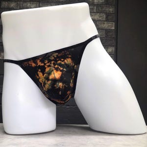 May include: Black underwear with a tie-dye pattern in shades of orange, yellow, and green. The underwear is displayed on a white mannequin. The background is a brick wall.