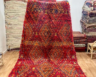 May include: A large, red rug with a geometric diamond pattern in orange, yellow, and blue. The rug has a thick, textured pile and a fringed edge. The rug is displayed in a room with other rugs and textiles.