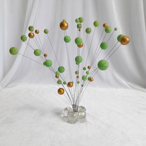 May include: A decorative floral arrangement featuring green and gold spheres of varying sizes on thin black stems. The spheres are arranged in a bouquet, held in a clear glass vase. The background is a white, draped fabric.