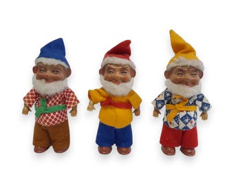 Puede incluir: Tres figuras de gnomos vintage con caras pintadas, barbas blancas y trajes coloridos. Los gnomos llevan sombreros azules, rojos y amarillos, y sus camisas tienen estampados de lunares, diseños florales y formas geométricas. Los gnomos están de pie.