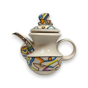 Könnte beinhalten: Eine cremefarbene Keramik-Teekanne mit einem lebendigen, mehrfarbigen geometrischen Muster und schwarzen Umrandungen. Das Design zeigt abstrakte Formen in Blau, Gelb, Orange und Violett. Sie hat einen passenden Deckel mit einem skulpturalen Knauf.
