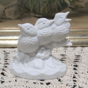 May include: White porcelain figurine of three birds perched on a branch. The birds are facing forward with their beaks open. The figurine is on a white base with a leafy design.