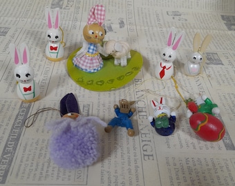 May include: A collection of wooden Easter bunny figurines, some with painted details, including a bunny wearing a blue and white dress, a bunny wearing a red bow tie, and a bunny holding a lamb. The figurines are arranged on a green and white base with a white background.