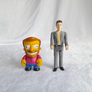 May include: Two small figurines, one is a yellow cartoon character with a pink shirt and blue pants, the other is a man in a gray suit and a beige shirt.
