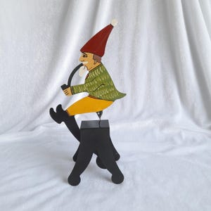 May include: A whimsical wooden figurine of a seated man smoking a pipe. The figure wears a red hat, green jacket, yellow pants, and black boots. The figurine is mounted on a black base with three legs, creating a unique decorative piece.