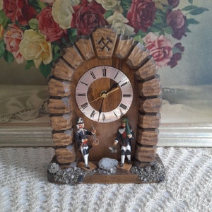May include: A wooden clock with a carved mountain scene and two figures of miners. The clock face is round with Roman numerals and a silver-toned hands. The clock is on a white lace tablecloth.