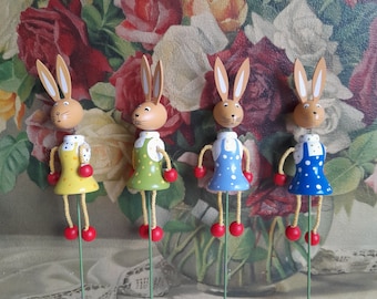 May include: Four wooden bunny figurines with painted details. Each bunny is wearing a different colored dress with white polka dots. The bunnies are on sticks and are arranged in a row.