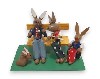 Peut inclure: Figurines en bois de lapins vêtus de différentes tenues, dont une veste bleue, un gilet rouge et un pantalon à pois. Un lapin est assis sur un banc, tandis que d'autres sont sur une base verte. Les figurines sont peintes en marron, bleu, rouge et blanc.
