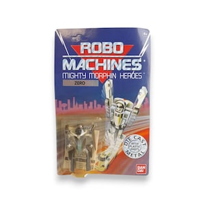 May include: A Robo Machines Mighty Morphin Heroes Zero action figure in its original packaging. The packaging is blue and white with the Robo Machines logo and the text "Die Cast Metal". The toy is a silver and black robot.