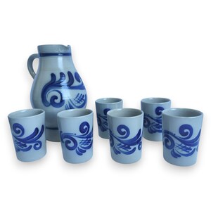 Op de afbeelding: Een lichtblauwe keramische kan met een handvat en tuit, en zes bijpassende bekers. Elk stuk heeft een donkerblauw, wervelend bloemmotief. De bekers zijn cilindrisch van vorm.