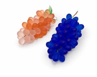 Puede incluir: Dos racimos decorativos de uvas hechos de vidrio esmerilado. Un racimo presenta tonos melocotón y naranja con una pequeña hoja de vidrio verde, mientras que el otro es de un azul profundo y vibrante con un tallo de alambre dorado en la parte superior.