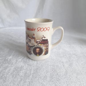 Könnte beinhalten: Weiße Keramiktasse mit einem roten und schwarzen Bild einer deutschen Stadtkulisse und dem Text "Weihnachtsmarkt 2009".