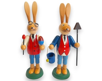 Puede incluir: Dos figuras de conejos de Pascua de madera. Un conejo lleva gafas y sostiene un huevo y una piruleta. El otro conejo sostiene un cubo y una herramienta de jardinería. Ambos conejos visten chalecos rojos, pantalones azules y tienen orejas largas.