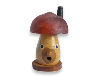 Puede incluir: Una figura de hongo de madera con una tapa roja y un tallo marrón claro. La figura tiene una expresión de sorpresa con ojos negros y una boca sorprendida. Una pequeña chimenea negra está en la parte superior del sombrero de hongo.